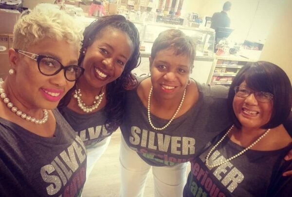 Four women, wearing matching 'SILVER' t-shirts and pearl necklaces, smile while standing together in a well-lit indoor setting near a counter.