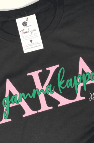 A black AKA Chapter Tee with pink Greek letters "AKA" and the text "gamma kappa" in green script. A tag attached to the shirt reads "Thank you for your purchase!".