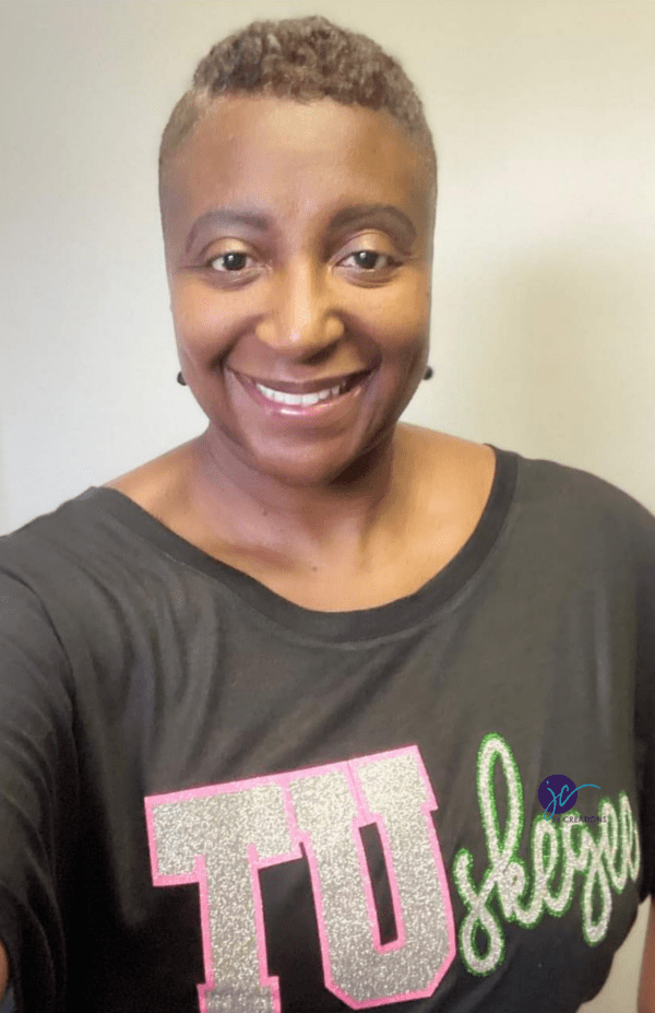 A smiling person wearing a black TUskegee Gamma Kappa Silver Star AKA Dolman Shirt with "Tuskegee" written on it in large, glittery letters poses for a photo.