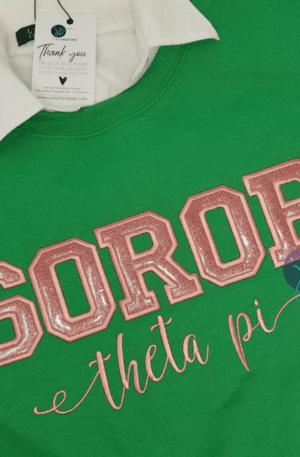A Pink SOROR Embroidered Unisex Sweatshirt with Chapter, featuring large pink glittery letters and "theta pi" in script font below, reminiscent of an Alpha Kappa Alpha embroidered soror shirt. A white collar and a tag are visible at the neckline.