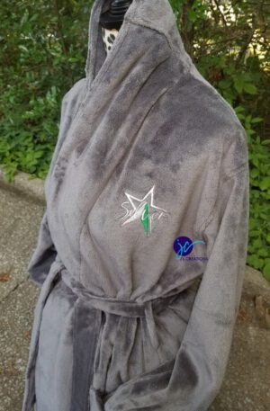 A mannequin displays a grey AKA Crest Plush Microfleece Shawl Collar Robe with "Star" and a logo embroidered on the left chest. The background includes greenery and a paved ground.