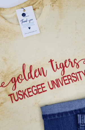 A t-shirt with "Golden Tigers Tuskegee University" in red text lays next to a pair of blue jeans. The light-colored Custom School/Mascot Embroidered Unisex Sweatshirt has a thank you tag attached.