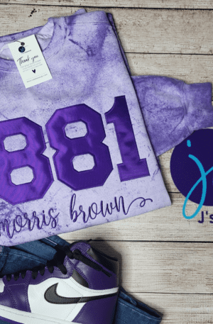 Purple sweater with "1881 Morris Brown" text, accompanied by a business card, a pair of jeans, and purple and white sneakers. The items are placed on a wooden surface beside a logo for JC's Creations, reminiscent of a Sigma Gamma Rho Soror Inspired Embroidered Unisex Sweatshirt collection.