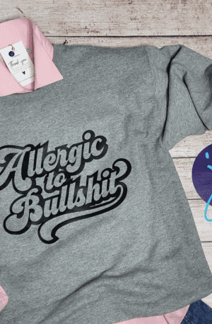 Gray sweatshirt with "Allergic to Bullshit (Black Design)" text, layered over a pink button-up shirt, lying on a wooden surface next to a "J's CREATIVE" logo.