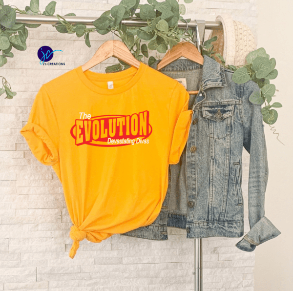 A yellow The Evolution Devastating Divas Unisex Tee with a tied hem is hanging on a clothing rack next to a denim jacket, both displayed against a white brick wall with green vines.