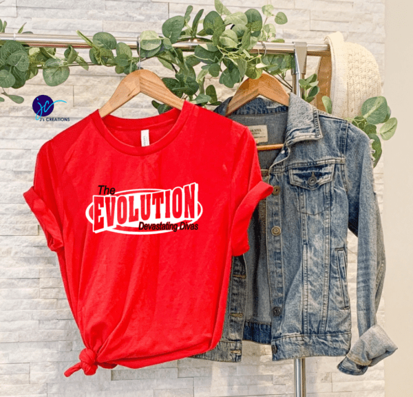 A red The Evolution Devastating Divas Unisex Tee with the text "The Evolution Devastating Divas" hangs next to a denim jacket. Both are on wooden hangers against a white textured background with a greenery decoration above.