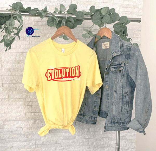 A yellow The Evolution Devastating Divas Unisex Tee with the text "The Evolution Demands Progress" is tied at the waist and displayed beside a blue denim jacket on a clothing rack with greenery in the background.