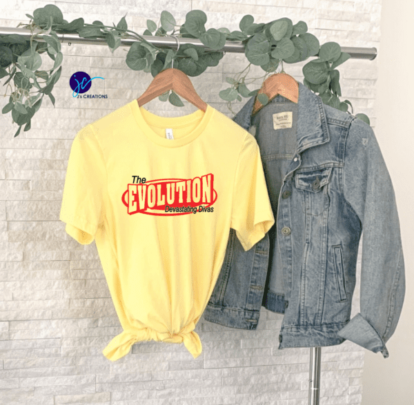 A yellow The Evolution Devastating Divas Unisex Tee is hanging next to a denim jacket on a white brick wall adorned with green leaves.
