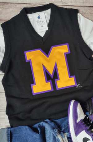 A Unisex Varsity Letter Sweater Vest with a large yellow "M" letter on the front is displayed on a wooden surface. It is placed over a white Alpha Kappa Alpha Soror Shirt next to a pair of purple and white sneakers and blue jeans.