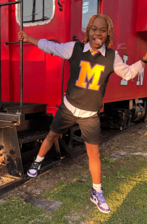A person with blonde hair poses playfully with one hand on a red train car and the other making a peace sign. They are wearing an Unisex Varsity Letter Sweater Vest, shorts, and an Alpha Kappa Alpha Soror Shirt underneath.
