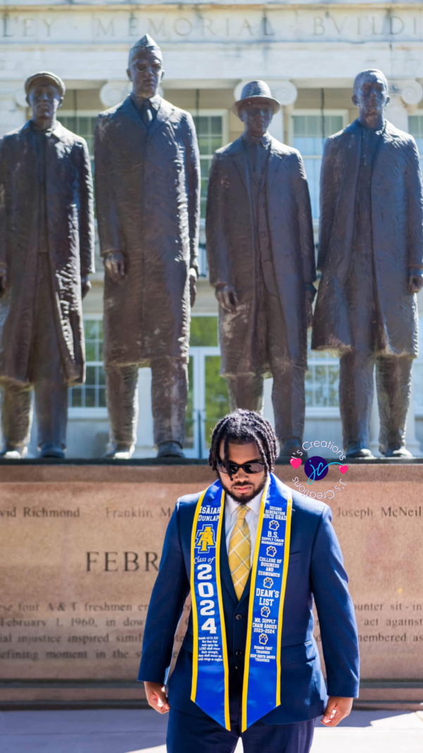 A person in a dark blue suit and Custom Graduation Stole stands in front of statues of four men. The background features a building facade with the words "Memorial Bldg" partially visible.