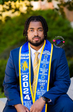 A graduate with a beard wearing a blue suit, gold tie, and Custom Graduation Stole that reads "Isaiah Dunlap, Class of 2024," sits outdoors. The stole includes honors and affiliations with North Carolina A&T State University.