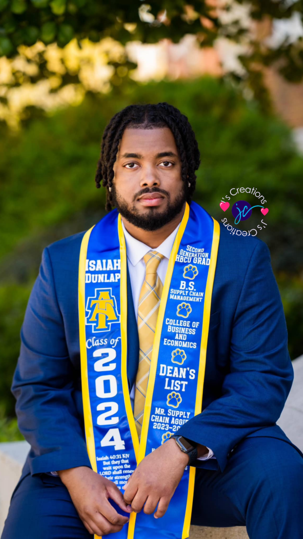 A graduate with a beard wearing a blue suit, gold tie, and Custom Graduation Stole that reads "Isaiah Dunlap, Class of 2024," sits outdoors. The stole includes honors and affiliations with North Carolina A&T State University.