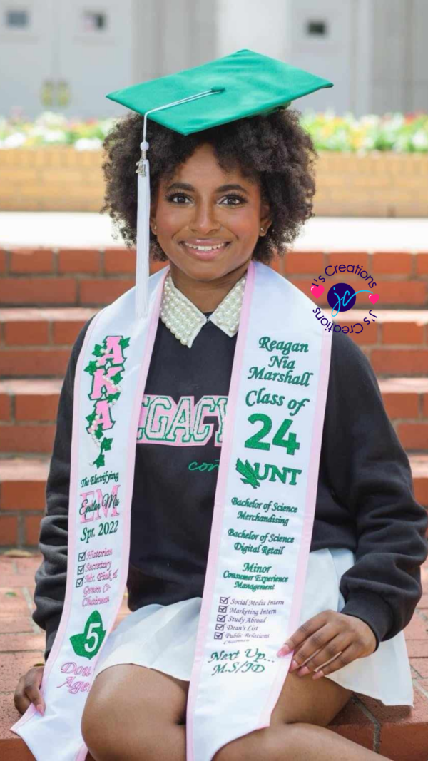 A woman in a graduation cap and gown sits on stairs, smiling. She wears a green cap and a Custom Graduation Stole with various text and symbols.